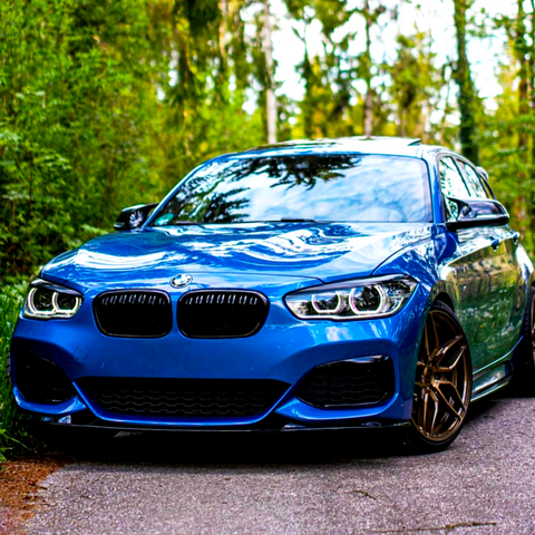 A blue car is parked on a forest road, surrounded by tall green trees and foliage, reflecting the sky on its shiny surface.