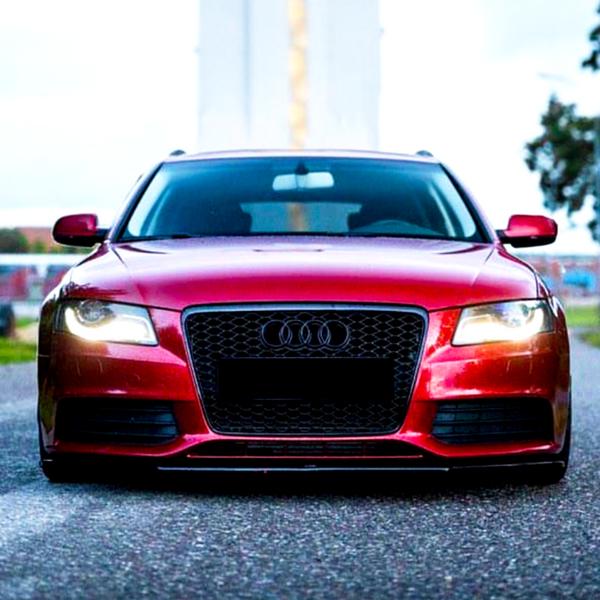 A red Audi car with illuminated headlights is stationary on a paved road, flanked by trees and buildings, under a cloudy sky.