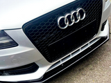 A silver car's front grill with a honeycomb pattern displays the Audi logo, consisting of four interlocking rings, against a concrete background.