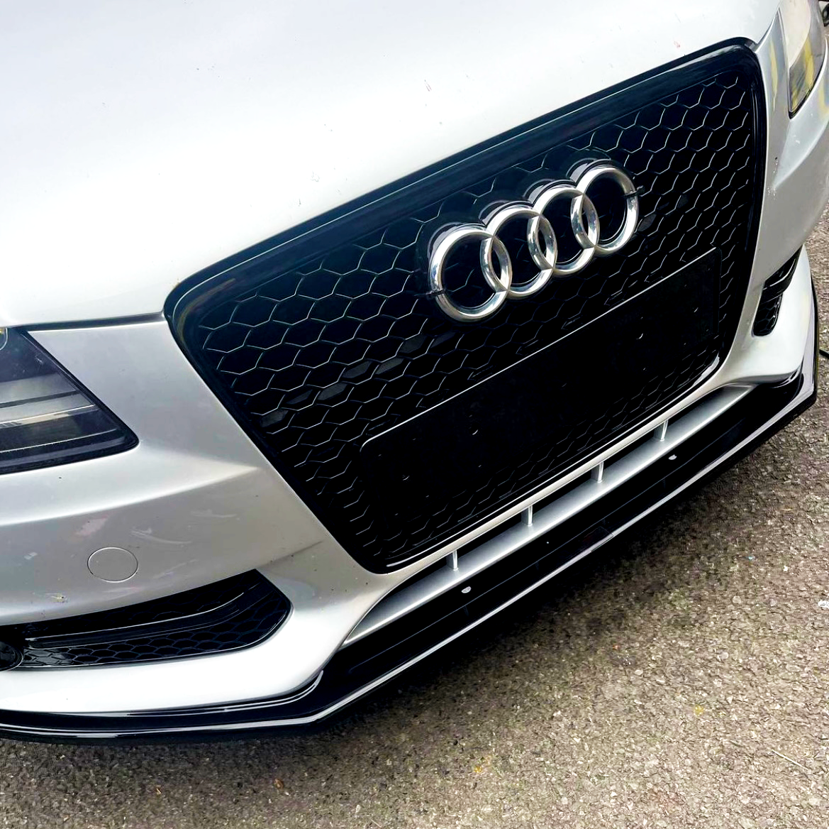 A silver car's front grill with a honeycomb pattern displays the Audi logo, consisting of four interlocking rings, against a concrete background.