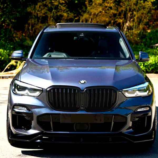 A dark-colored car is parked on a sunlit road, surrounded by lush green foliage. The car's front features a distinctive grille and sleek headlights.