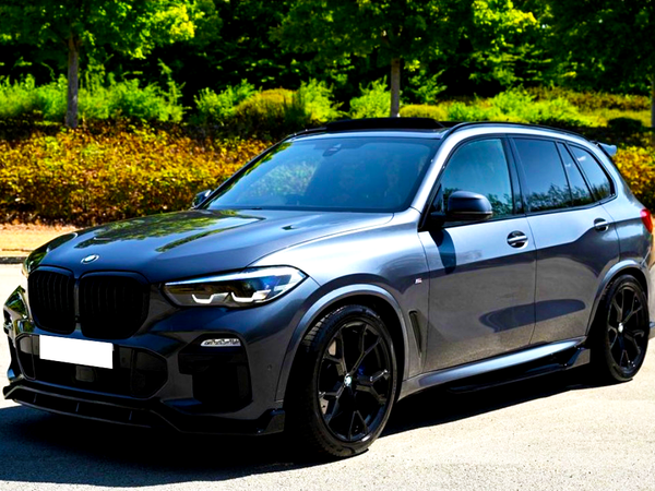 A sleek, dark-colored SUV is parked on a sunlit road, surrounded by lush green trees and bushes in the background.