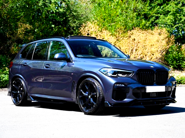 A sleek, dark-colored SUV is parked on a paved surface, surrounded by lush green trees and bushes in the background, with natural sunlight casting shadows.