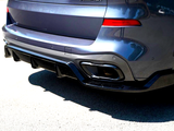 A dark metallic car's rear end, featuring dual exhaust tips and a diffuser, is parked on a sunlit asphalt surface. The vehicle's body reflects nearby buildings and trees.