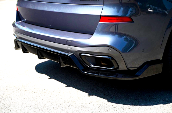 A dark metallic car's rear end, featuring dual exhaust tips and a diffuser, is parked on a sunlit asphalt surface. The vehicle's body reflects nearby buildings and trees.