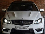 A white Mercedes-Benz car with illuminated headlights is parked in front of a brick wall. The license plate displays the text 