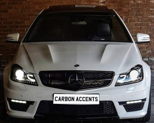A white Mercedes-Benz car with illuminated headlights is parked in front of a brick wall. The license plate displays the text "CARBON ACCENTS."