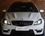 A white Mercedes-Benz car with illuminated headlights is parked in front of a brick wall. The license plate displays the text "CARBON ACCENTS."