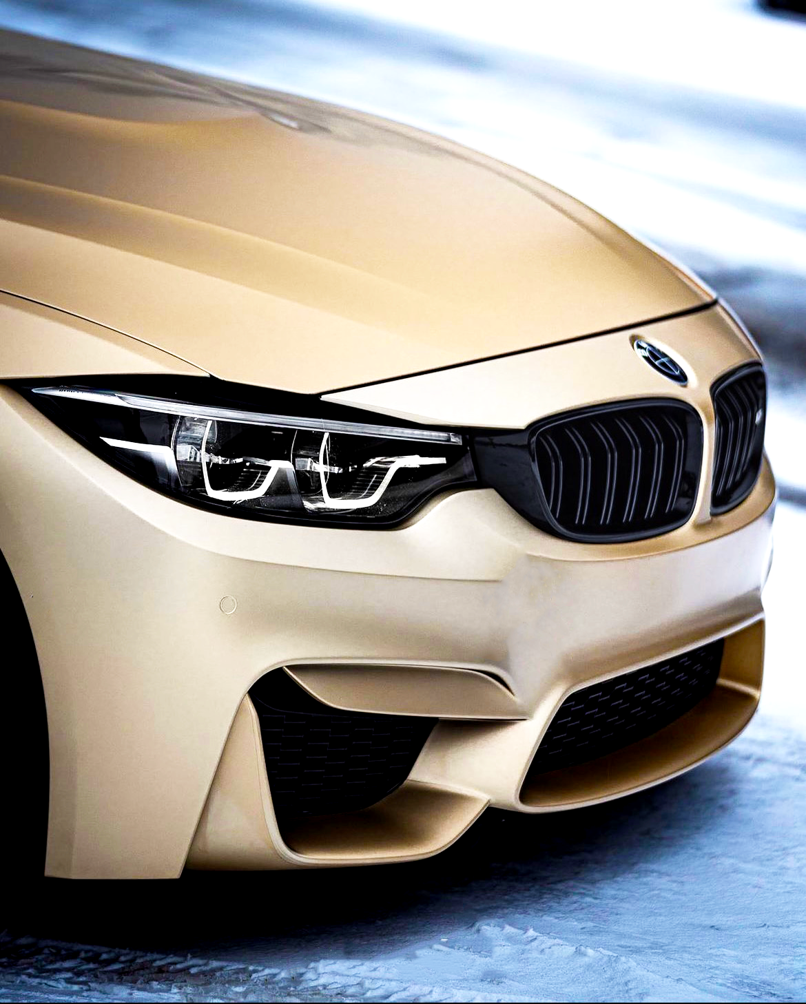 A gold BMW car with sleek front headlights and grille, parked on a snowy ground, showcasing its elegant design and smooth finish.