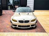 A white BMW car sits parked on a brick driveway in front of a modern building, featuring yellow-tinted headlights and a sleek, aggressive front design.