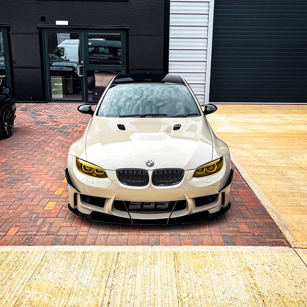 A white BMW car sits parked on a brick driveway in front of a modern building, featuring yellow-tinted headlights and a sleek, aggressive front design.