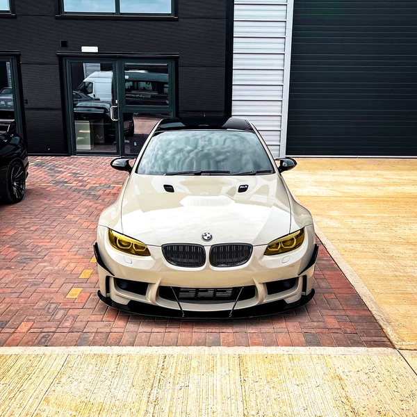 A white car with yellow-tinted headlights is parked on a brick driveway in front of a modern building with large windows and a garage door.