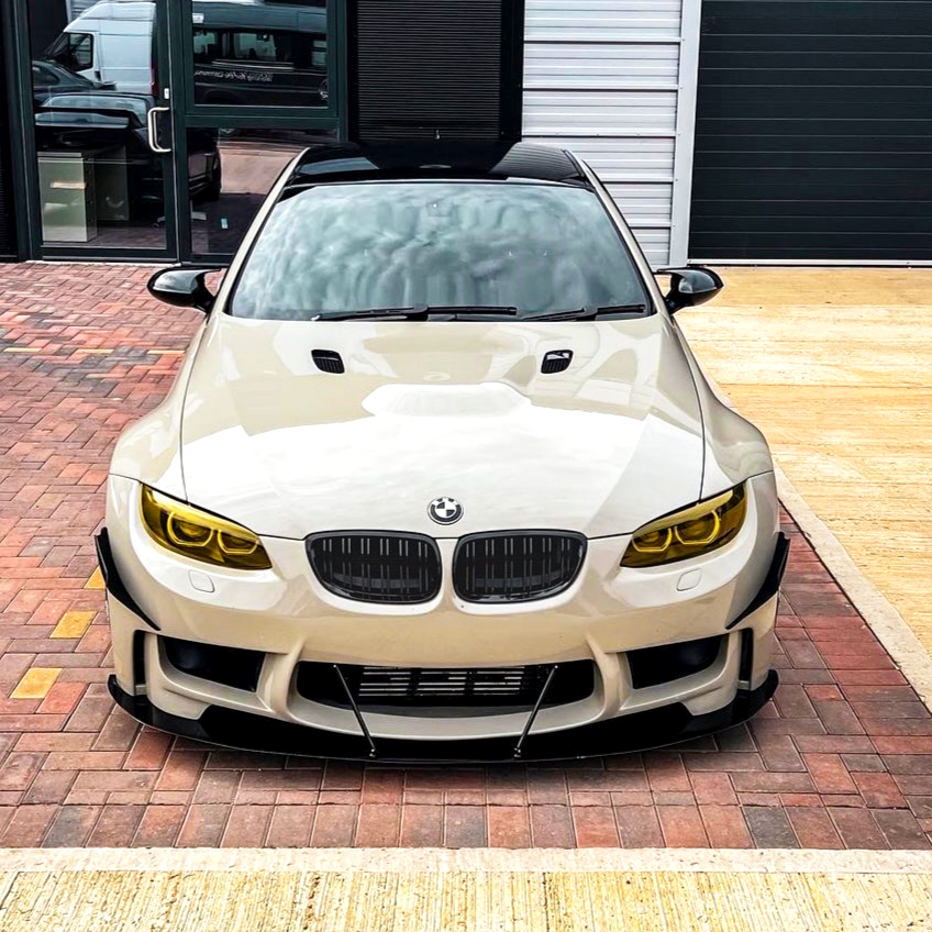 A modified white car with yellow-tinted headlights and aggressive front bumper stands parked on a brick driveway, in front of a modern building with large windows and dark doors.