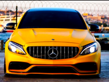 Yellow Mercedes-Benz car is parked, facing forward with headlights on, in front of a wire fence. The background includes a blurred water scene and distant buildings.