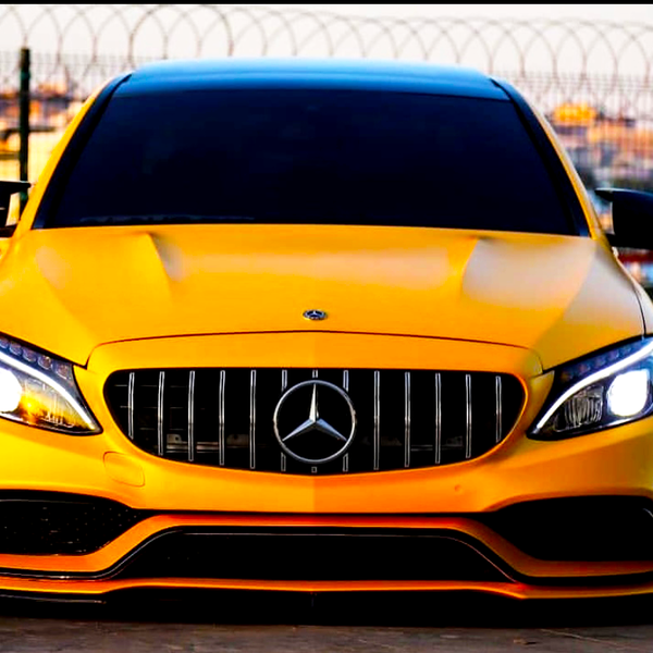 Yellow Mercedes-Benz car is parked, facing forward with headlights on, in front of a wire fence. The background includes a blurred water scene and distant buildings.