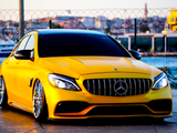 A yellow Mercedes-Benz car is parked on a paved surface beside a waterfront, featuring a distinctive grille and large chrome wheels, with a cityscape visible in the background.