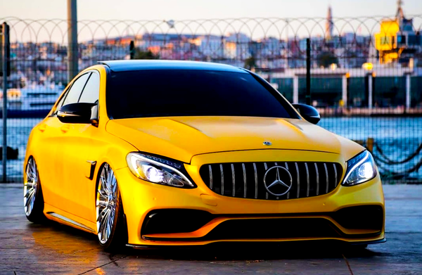 A yellow Mercedes-Benz car is parked on a paved surface beside a waterfront, featuring a distinctive grille and large chrome wheels, with a cityscape visible in the background.