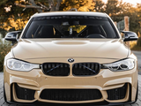 A tan BMW car is parked on a paved surface, facing forward. Trees with golden leaves surround the scene. The windshield displays the text 