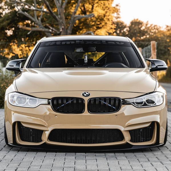 A tan BMW car is parked on a paved surface, facing forward. Trees with golden leaves surround the scene. The windshield displays the text "#tmhxclsv."