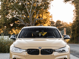 A beige BMW car is parked on a cobblestone driveway with trees in the background. The windshield displays the text 