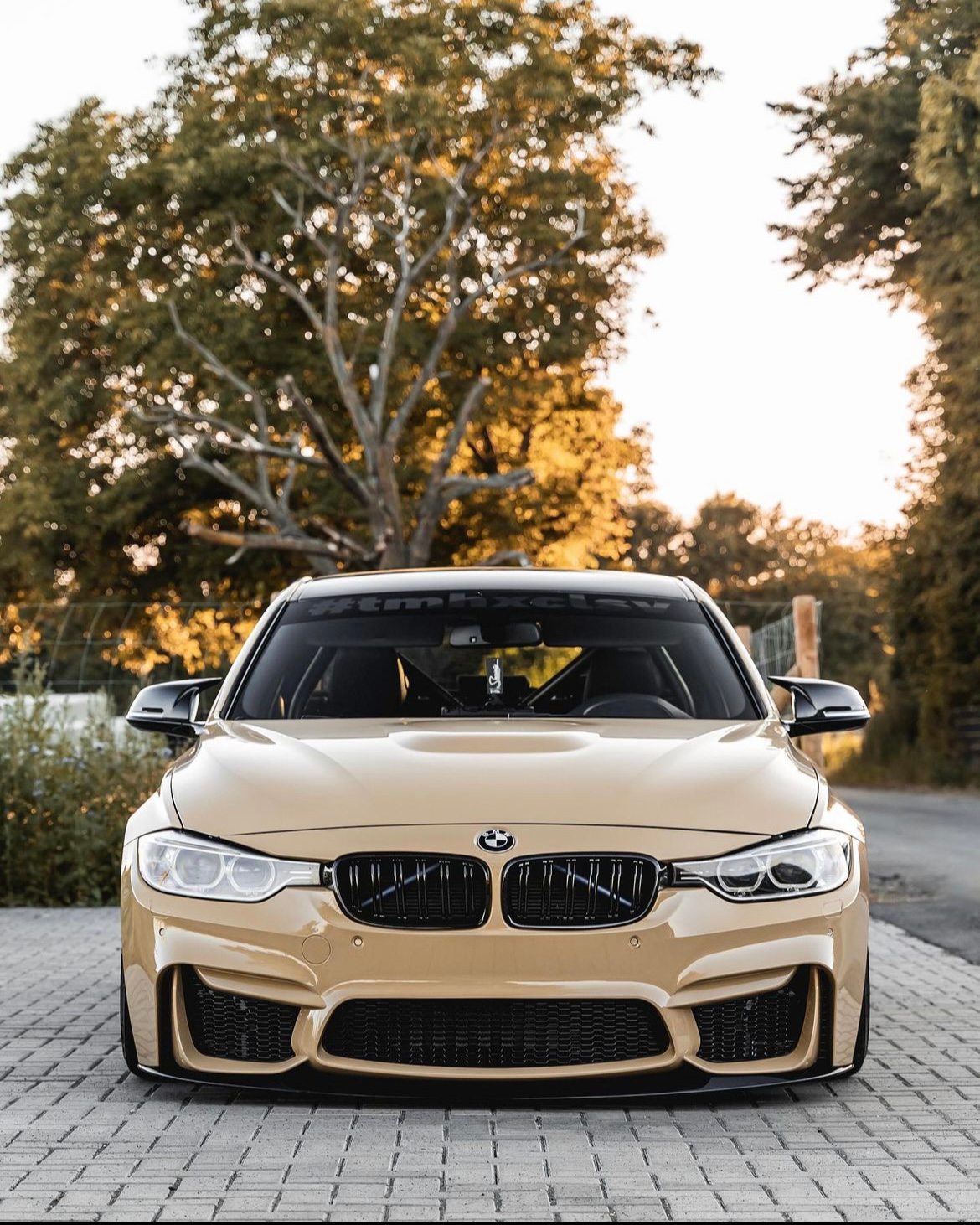 A beige BMW car is parked on a cobblestone driveway with trees in the background. The windshield displays the text "#tmhxct".