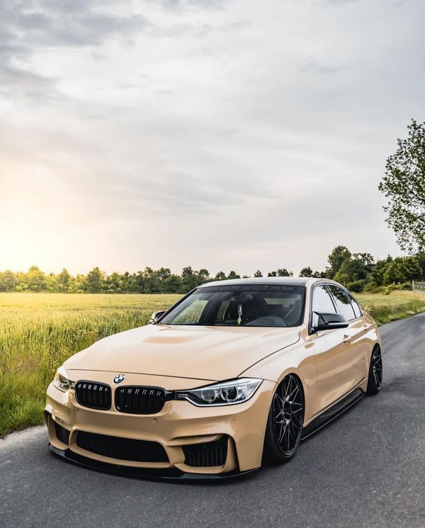 A beige sports car is parked on a paved road, surrounded by green fields and trees, under a cloudy sky, with sunlight casting a warm glow.