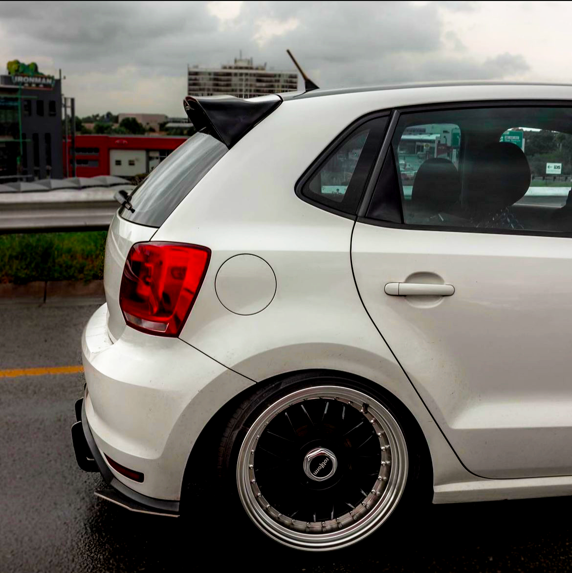 A white car with custom wheels is parked on a wet road near a cityscape. The background features buildings and cloudy skies, creating an urban atmosphere.