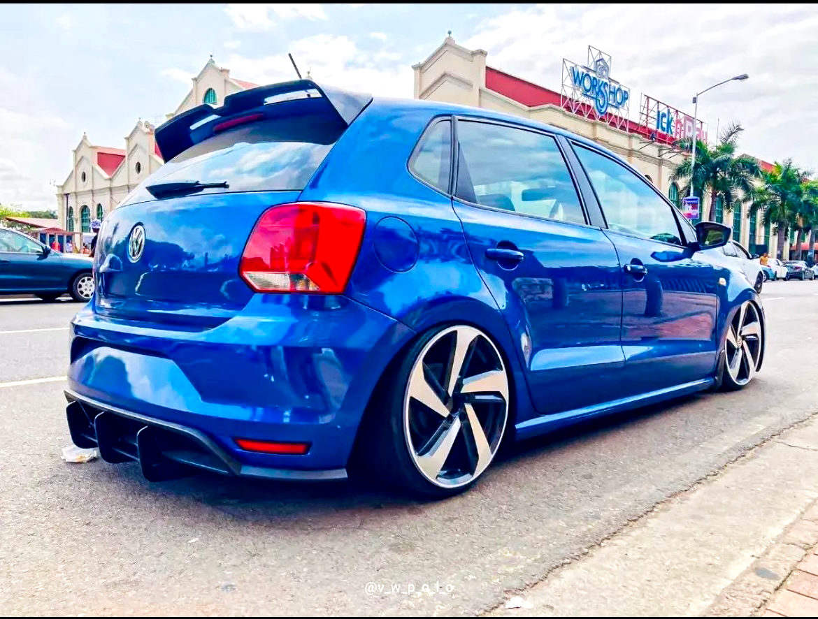 A blue Volkswagen hatchback with aftermarket rims is parked on the street. It features tinted windows, and a rear spoiler, with a building and palm trees in the background. Text: "@v_w_p_olo."