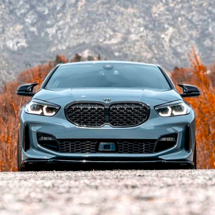 A sleek car, viewed from the front, is parked on a gravel surface. The vehicle has distinctive headlights and a prominent grille. In the background, there are autumnal trees and mountains.