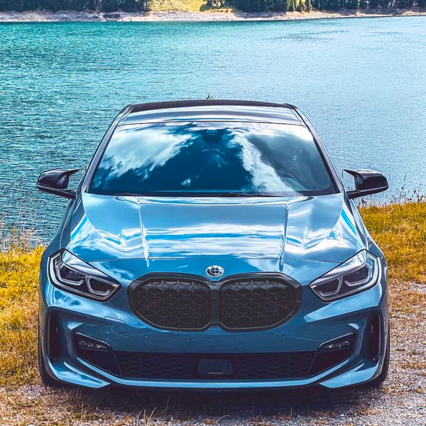 A sleek blue car is parked on grass beside a calm, blue lake, reflecting clouds. Trees are visible in the distant background.