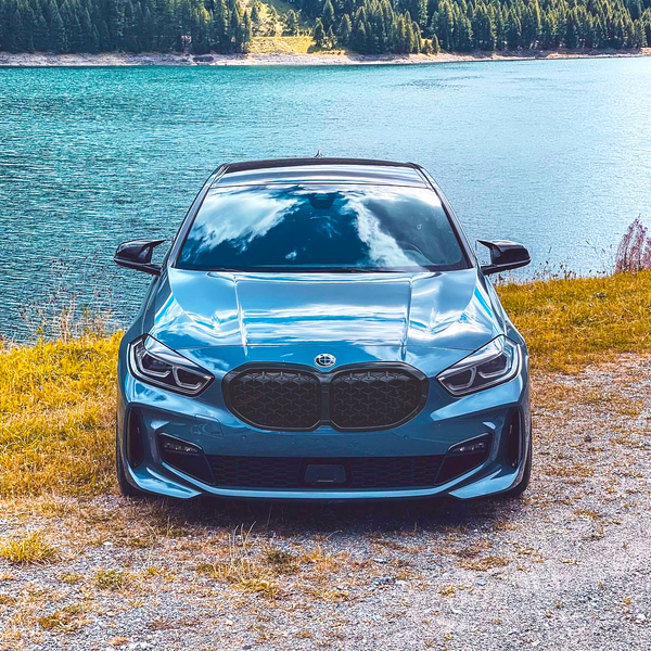 A blue car is parked on gravel near a lake, reflecting clouds. Behind, dense green forest rises over hillsides, contributing to a serene, natural landscape.