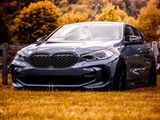 A sleek gray car is parked on vibrant autumn grass, bordered by a wooden fence. Behind, trees in warm fall colors and hills create a scenic backdrop.