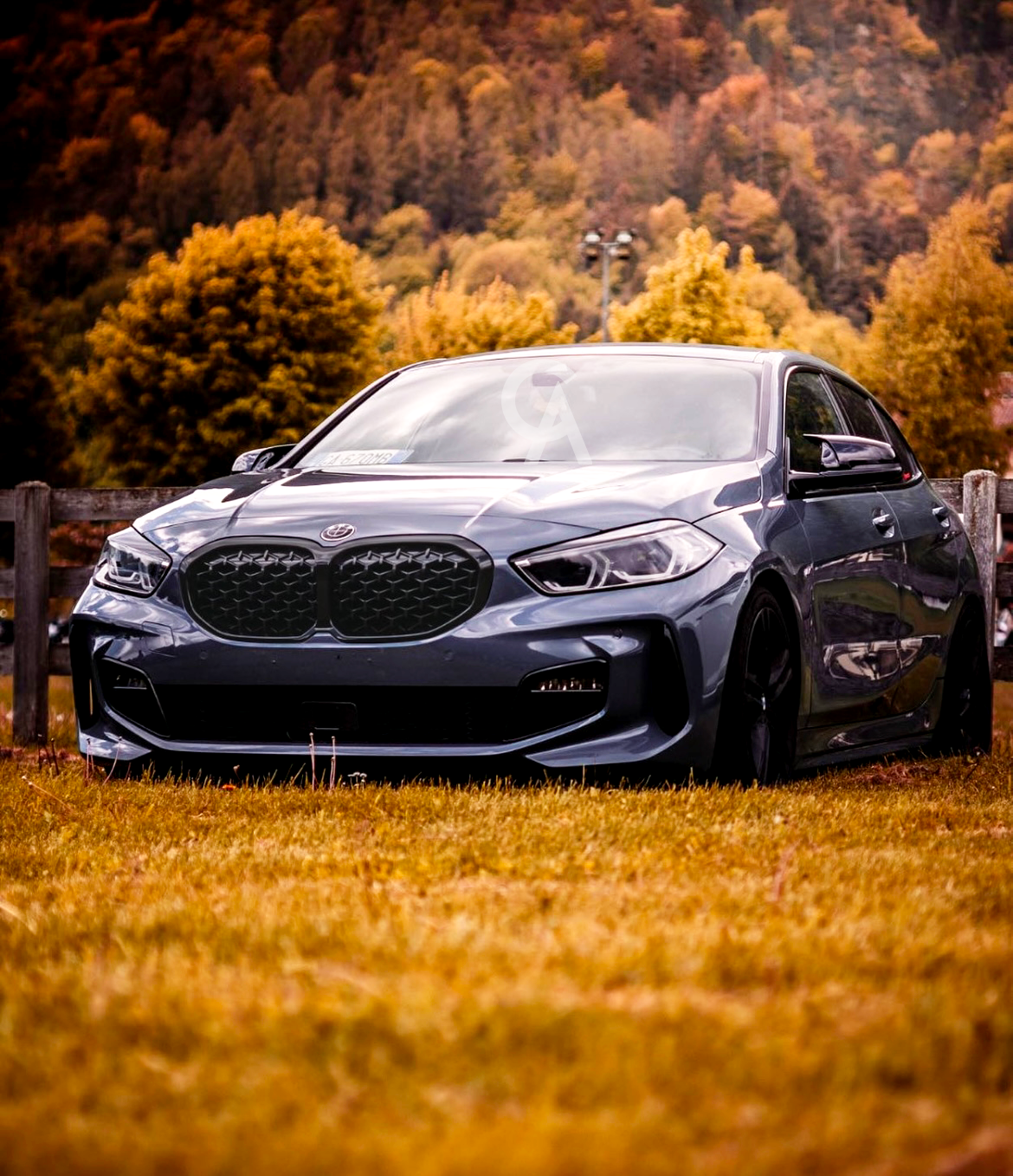 A sleek gray car is parked on vibrant autumn grass, bordered by a wooden fence. Behind, trees in warm fall colors and hills create a scenic backdrop.