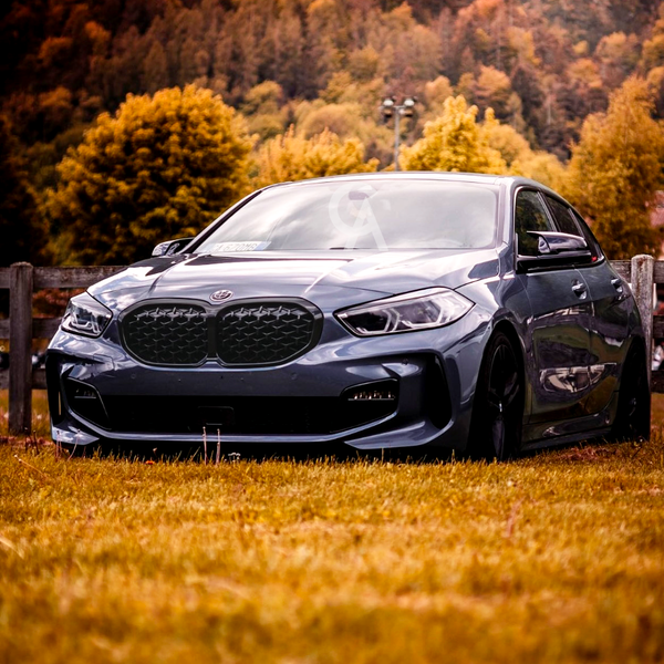 A sleek, blue car is parked on yellow grass, surrounded by a wooden fence, with a backdrop of autumn trees and hills. A windshield sticker reads "ALPINE."