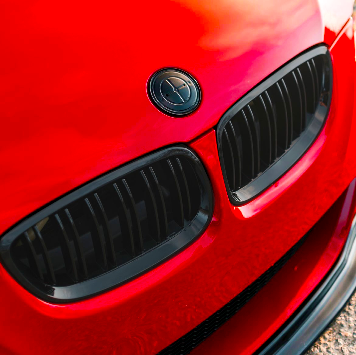 Red car with two black kidney grilles on the front, parked on an outdoor surface. The car's glossy finish reflects the sunlight.