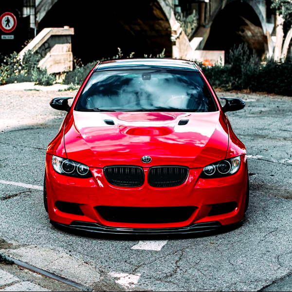 A shiny red sports car with a sleek design is parked on a cracked pavement, under a bridge with greenery surrounding it. A circular road sign is visible in the background.