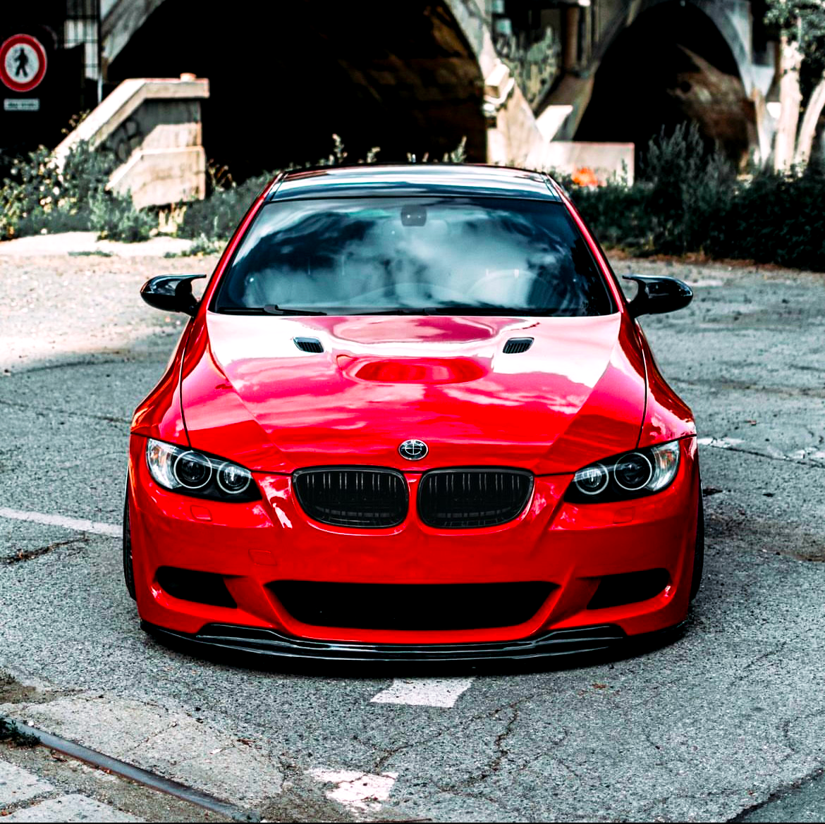 A red sports car is parked on a cracked pavement with a bridge and greenery in the background. A "no entry" sign is visible on the left side.