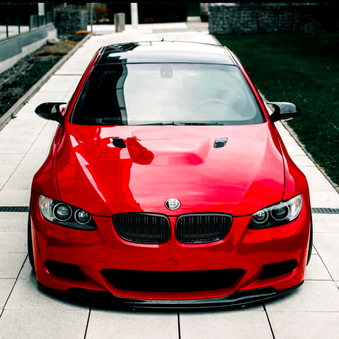 A red sports car, parked on a paved area, reflects the surroundings on its shiny surface. The environment includes a walkway and a patch of grass on the side.