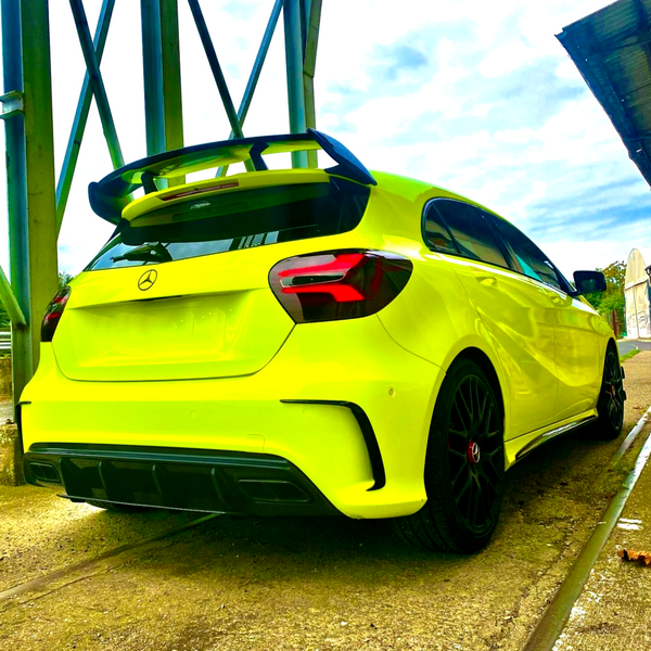 A bright yellow Mercedes-Benz hatchback is parked on a concrete surface, featuring a prominent rear spoiler and black wheels, under a metal structure on a partly cloudy day.