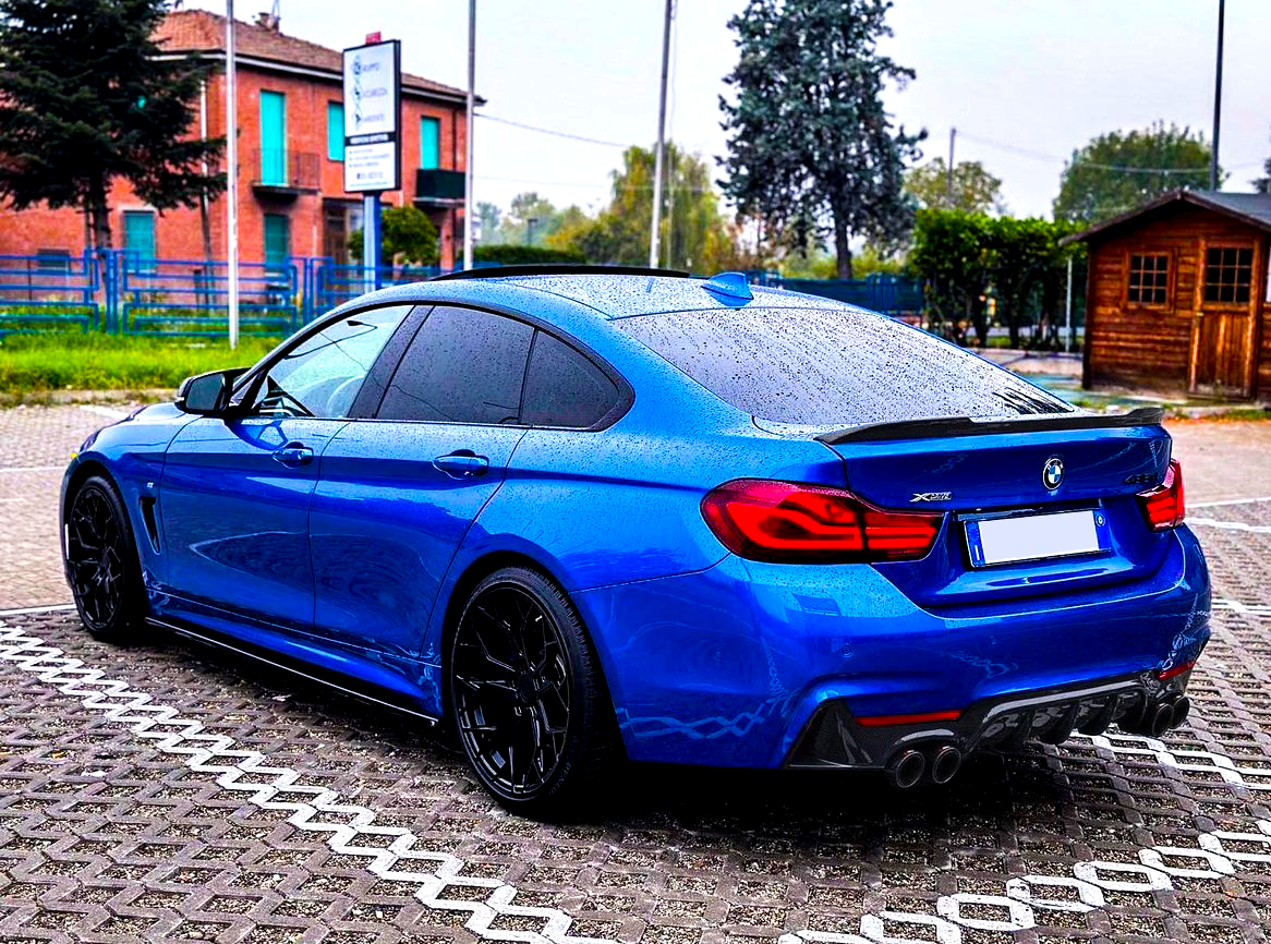 Blue BMW car parked, covered in raindrops, in a brick-paved lot with a wooden shed and trees in the background. Text includes “BMW,” "X Drive," and "420i".