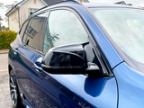 A blue car is parked on a wet driveway, showing a side mirror and window. It reflects nearby houses and a cloudy sky in a suburban neighborhood.