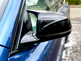A side mirror, sleek and black, reflects nearby houses and streetlights. It is attached to a shiny blue car parked on a wet driveway in a suburban neighborhood.