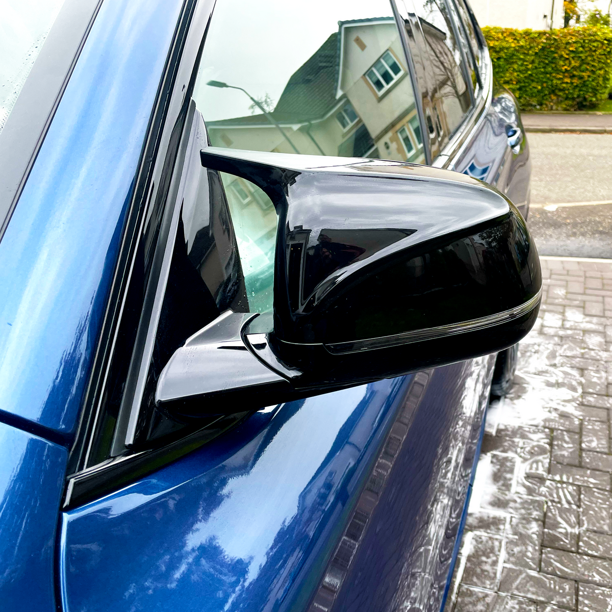 A side mirror, sleek and black, reflects nearby houses and streetlights. It is attached to a shiny blue car parked on a wet driveway in a suburban neighborhood.