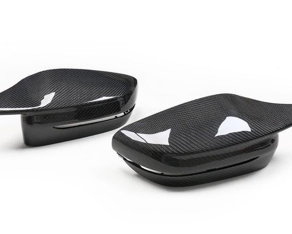 Two carbon fiber side mirror covers rest on a white background, showcasing a sleek, glossy finish with a subtle, woven pattern and reflection details.