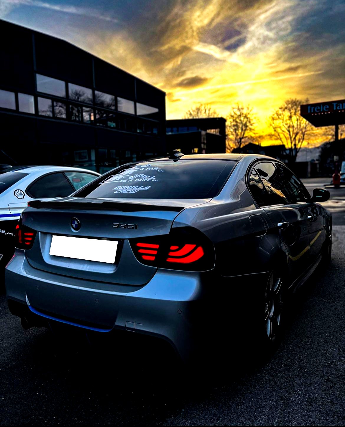 A silver BMW 335i with illuminated rear lights is parked near other vehicles at sunset. The rear window displays the text: "Good life, I say... drive like a maniac... Gorilla Gang."