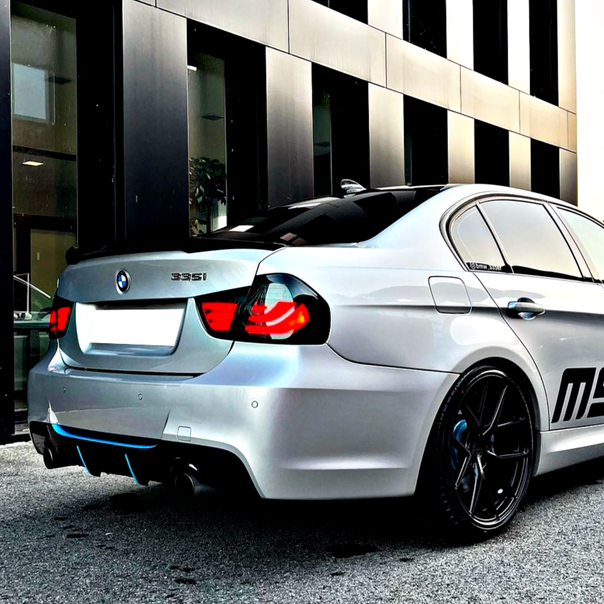 A white BMW 335i is parked on a street, displaying black rims and partially lit taillights. It's positioned in front of a modern building with large windows.