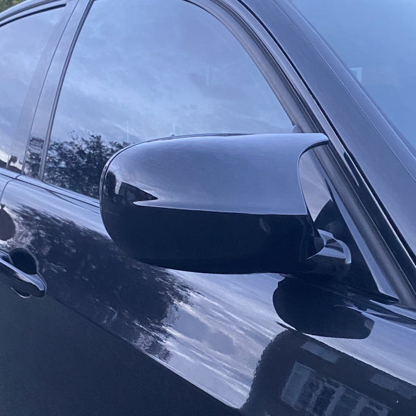 A car's side mirror reflects the surroundings, attached to a sleek, dark vehicle door. The background shows trees and sky, reflecting on the tinted windows and shiny surface.