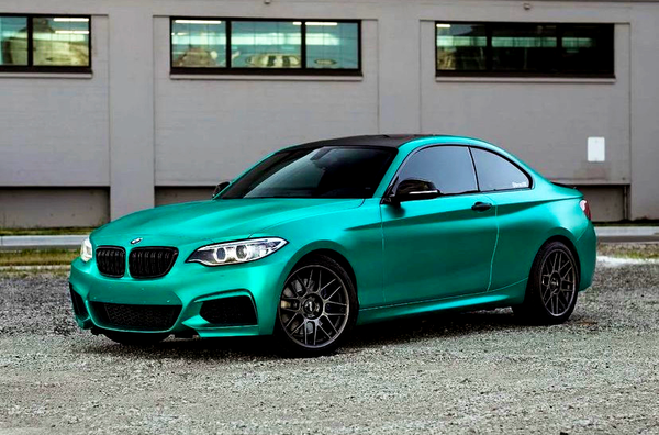 A teal sporty car is parked on a gravel surface against a backdrop of a building with several windows.