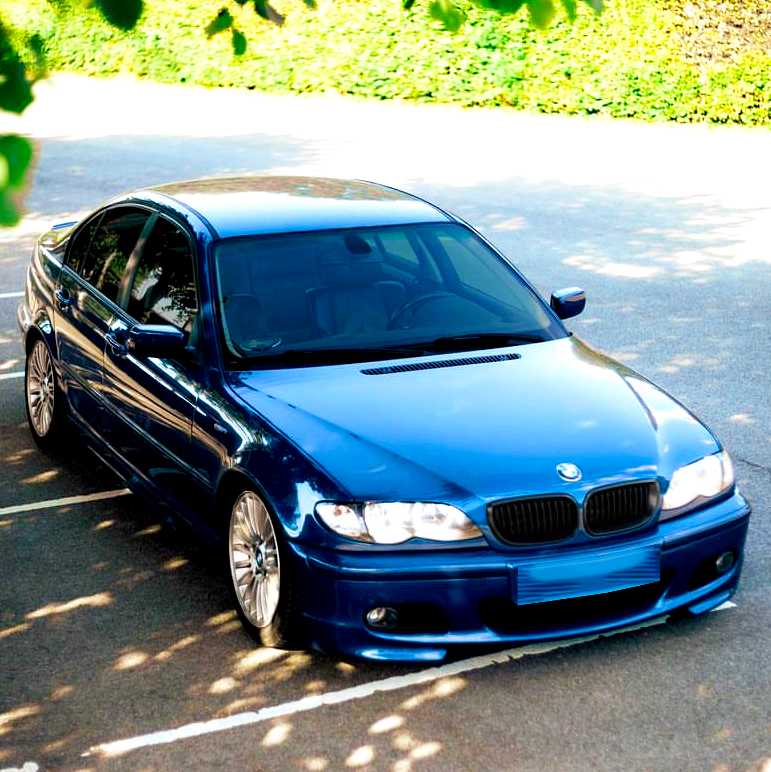 Blue sedan parked in a sunlit lot, surrounded by greenery. Bright reflections highlight the car's glossy finish, while tree shadows partially cover the asphalt.