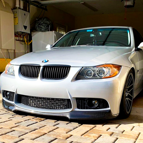A silver BMW car is parked in a garage with a partially visible water heater. Sunlight casts shadows across the vehicle, highlighting its sleek design on a paved surface.